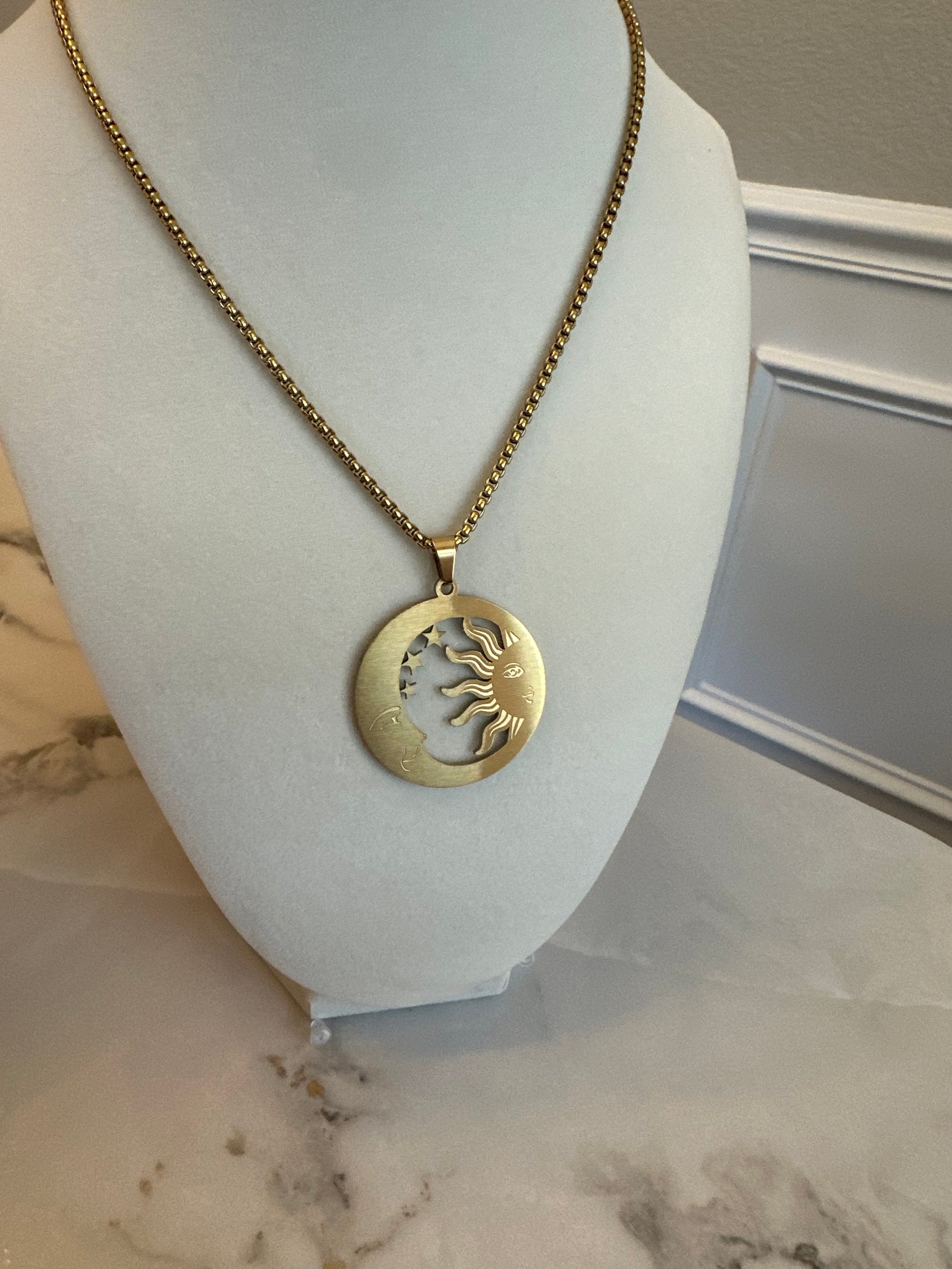 Gold necklace with a sun pendant on a mannequin bust against a marble wall.