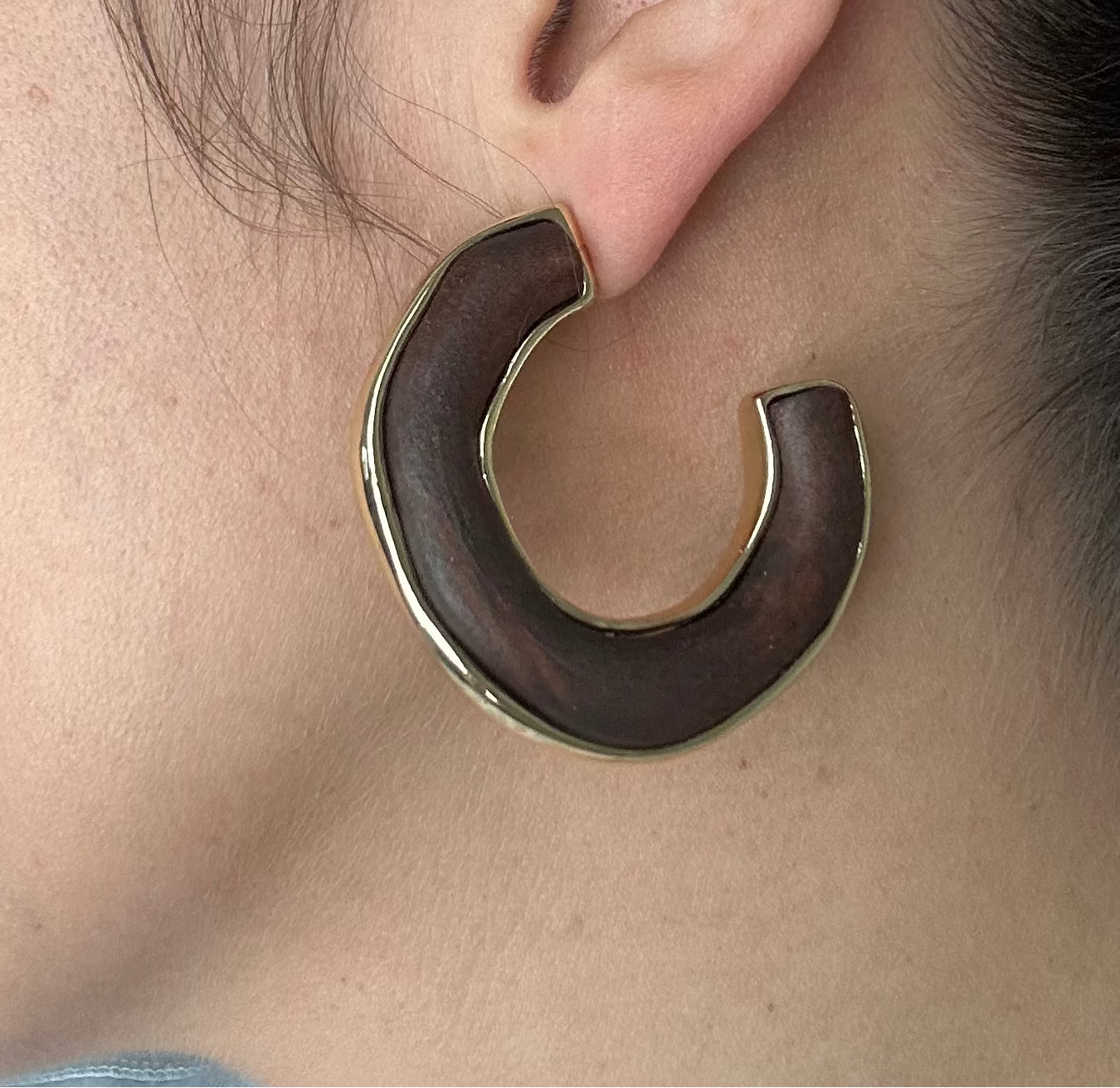 A close-up side profile of a woman wearing a large, chunky C-shaped wooden hoop earring with a polished gold-toned metal rim.