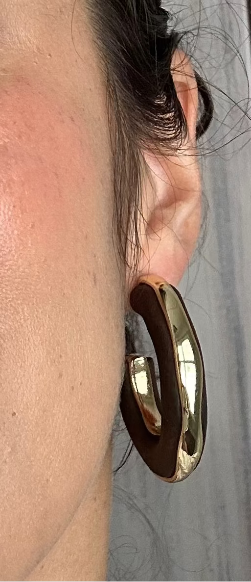 A close-up selfie of a woman wearing large, chunky gold hoop earrings and a light gray cowl-neck top, with a neutral indoor background.