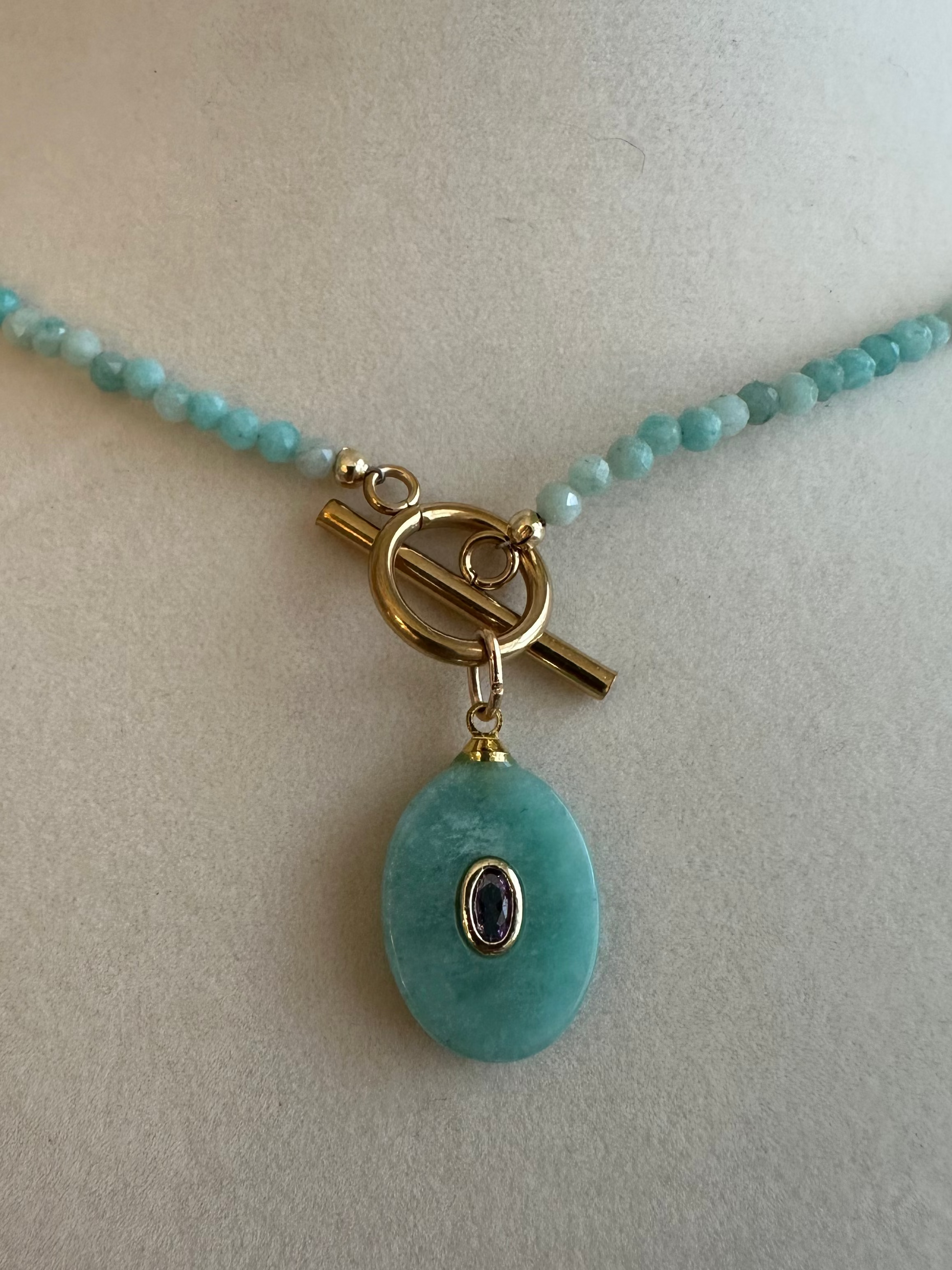 A turquoise beaded necklace with a large oval turquoise pendant and a gold toggle clasp, displayed on a cream-colored mannequin bust.
