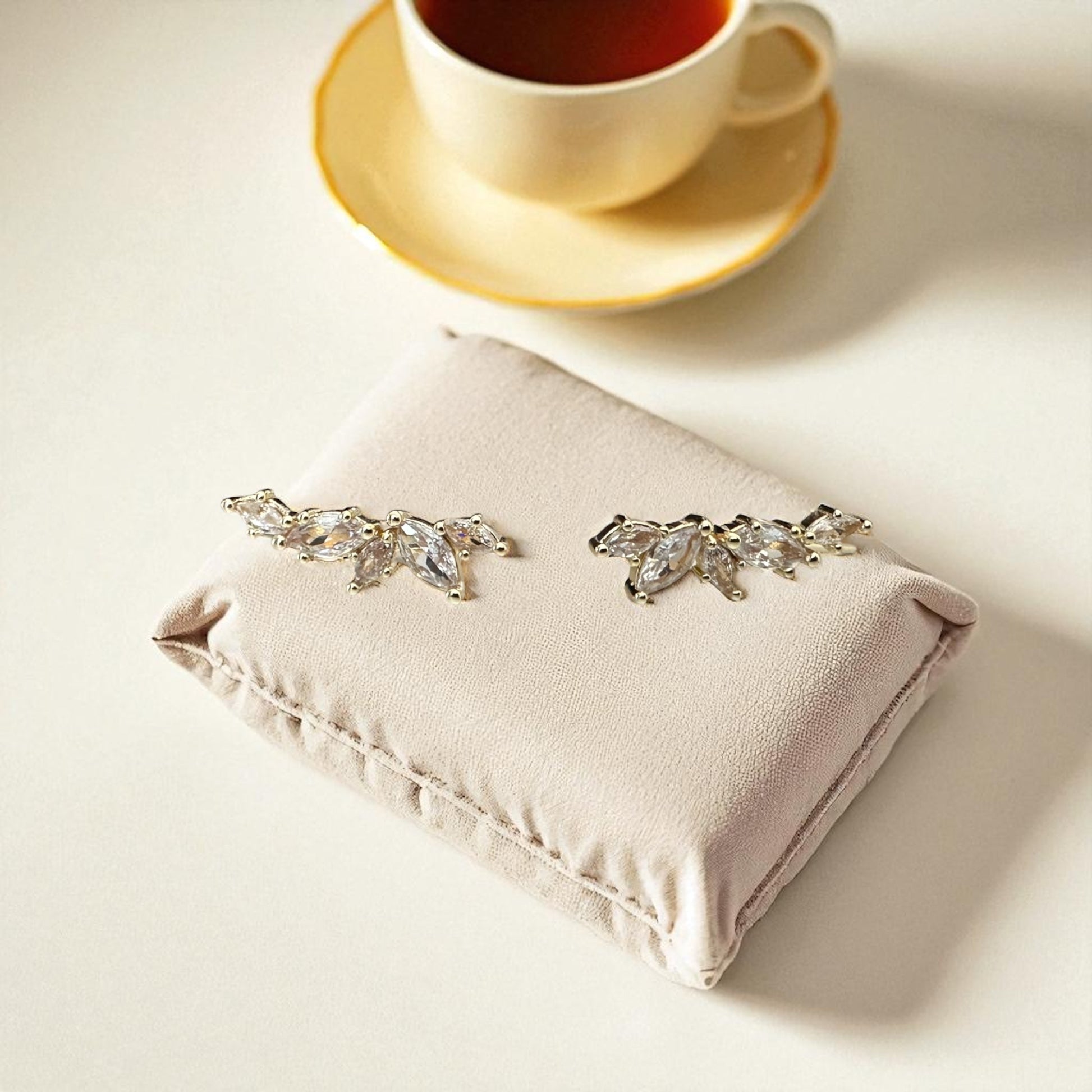 Pair of silver leaf-shaped earrings on a pink cushion with a cup of tea in the background.
